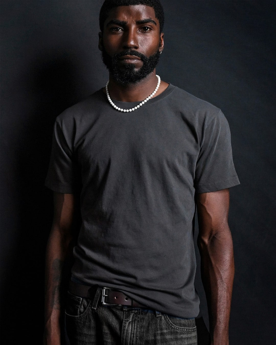 Man in a white t-shirt and necklace, standing against a dark background.