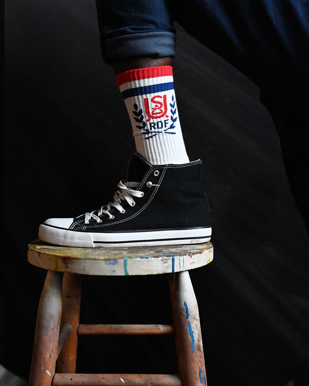 Foot in a black canvas sneaker and white socks resting on a painted wooden stool.