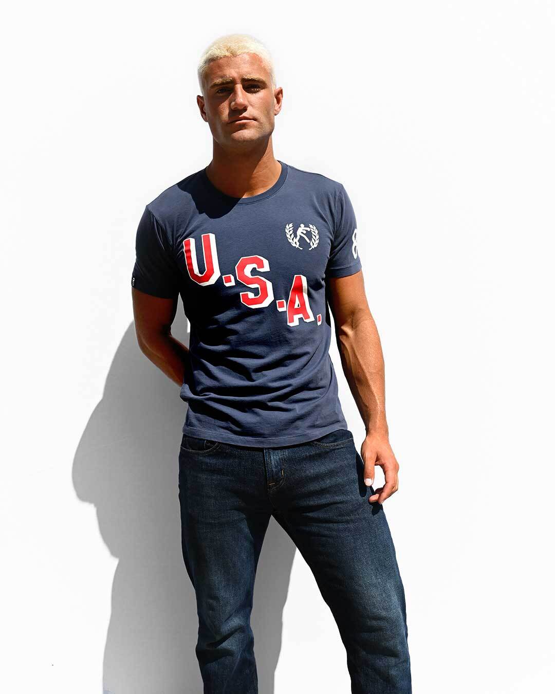 Man with blonde hair wearing a navy 'U.S.A.' shirt and jeans stands against a plain white background.